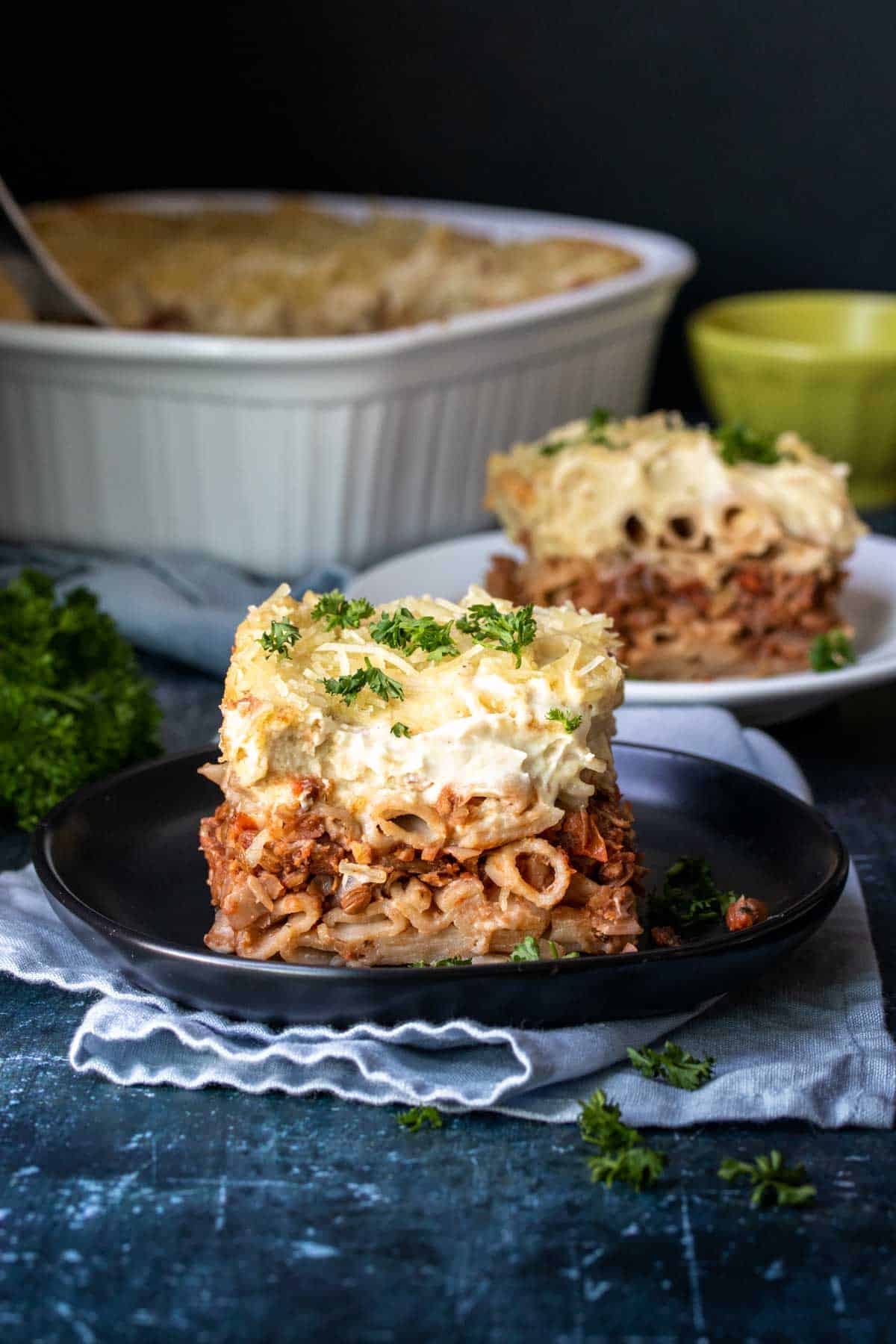 Front view of a piece of pastitsio on a black plate sprinkled with parsley and more pastitsio in the background