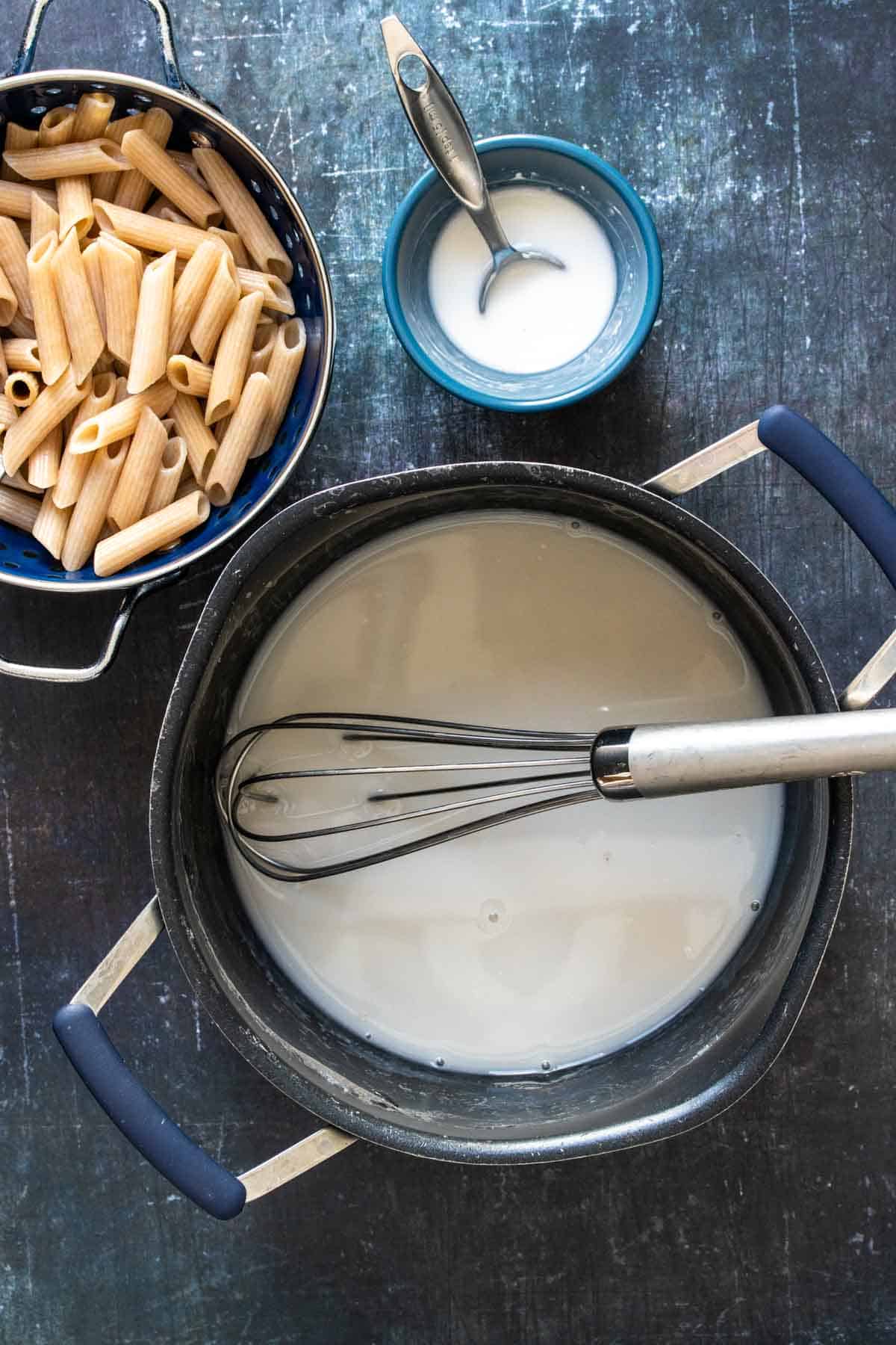 Top view of a large pot with white liquid and a whisk inside next to pasta and another white liquid