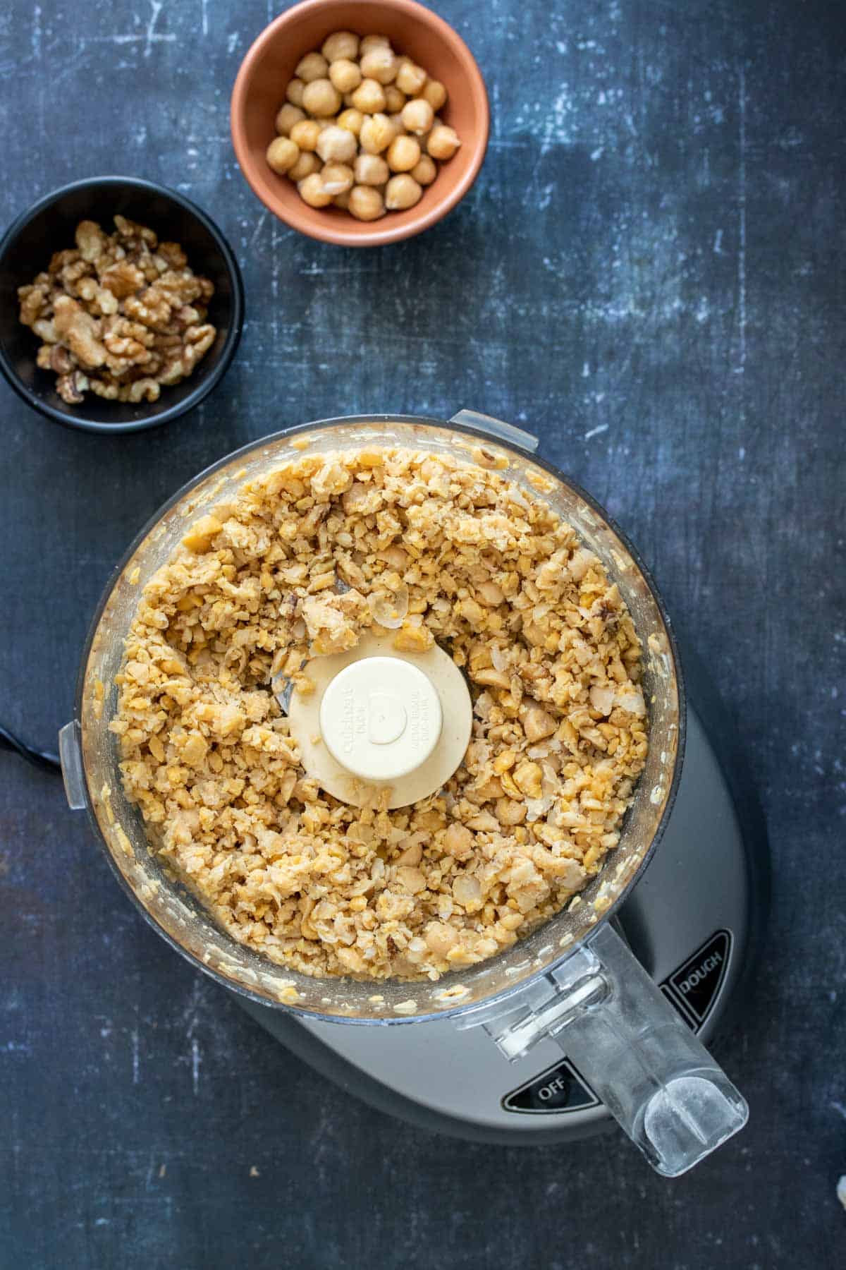 Top view of a food processor with chopped chickpeas and walnuts next to bowls of whole ones