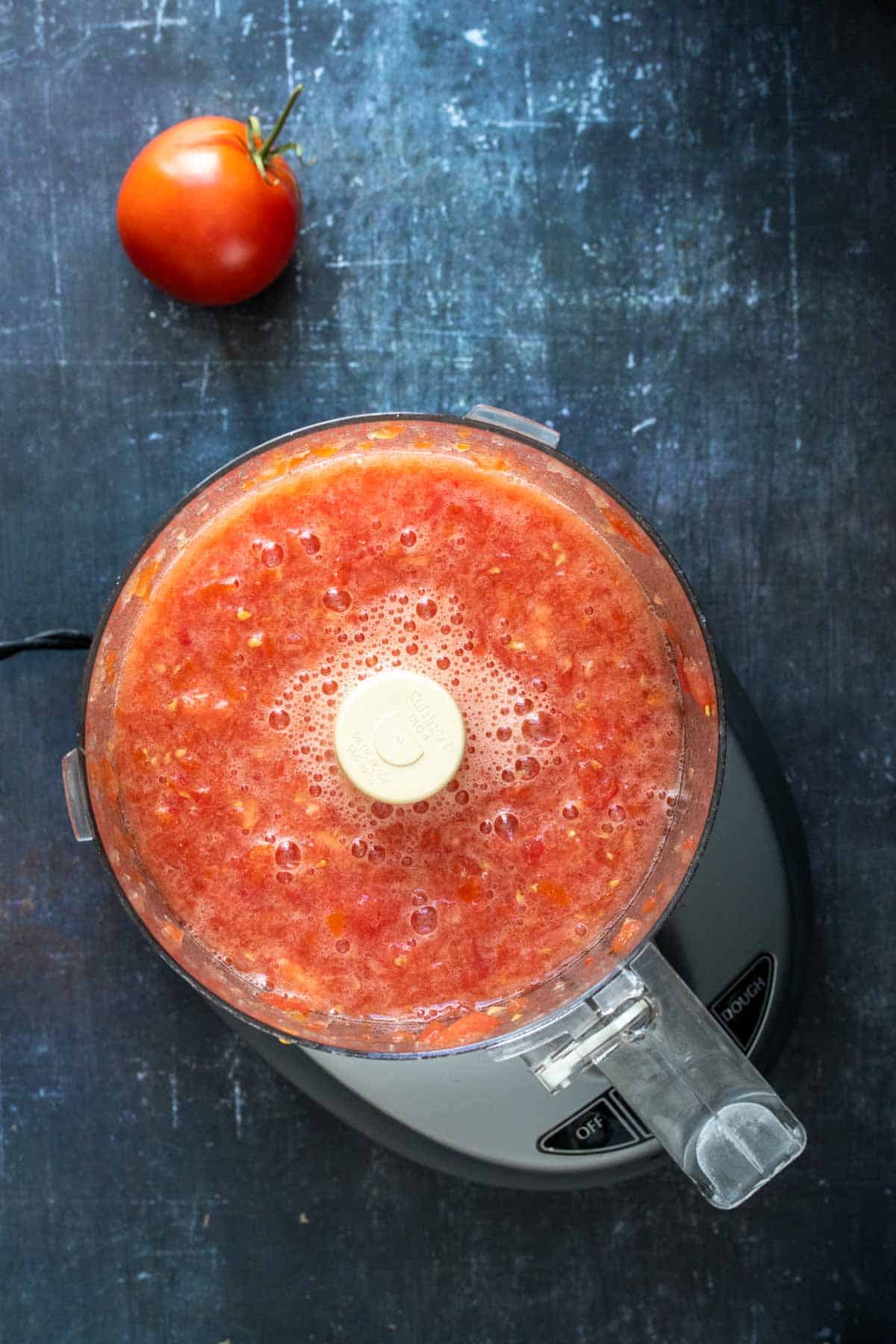 Top view of a food processor with chopped tomatoes next to a whole tomato