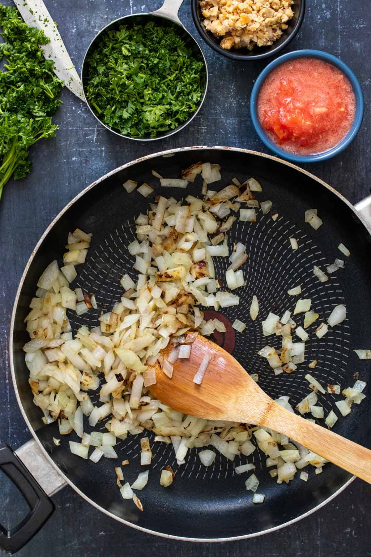 Top view of a pan with chopped onions being mixed by a wooden spoon next to bowls of herbs, tomatoes and chopped chickpeas