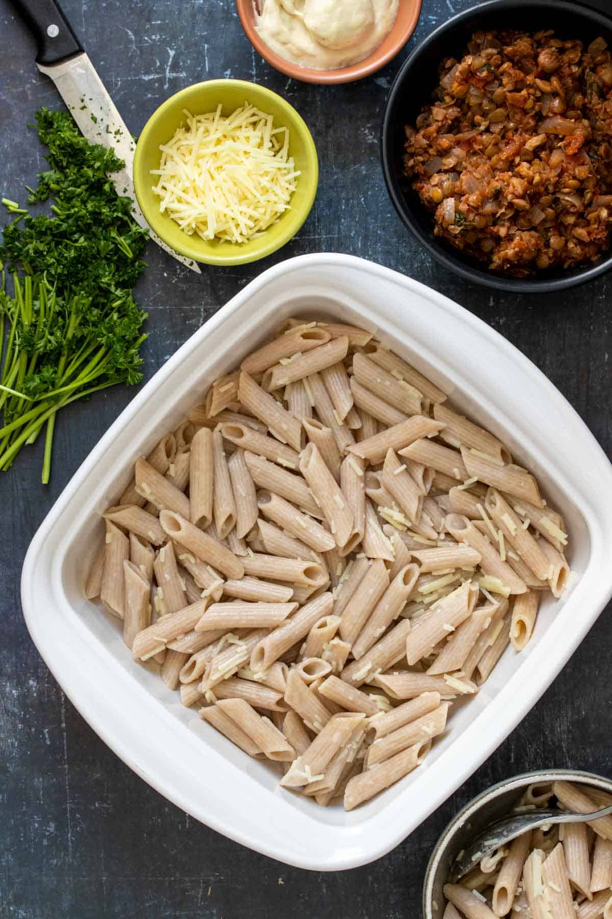 Top view of a white baking dish filled with penne pasta next to other ingredients needed to make Pastitsio