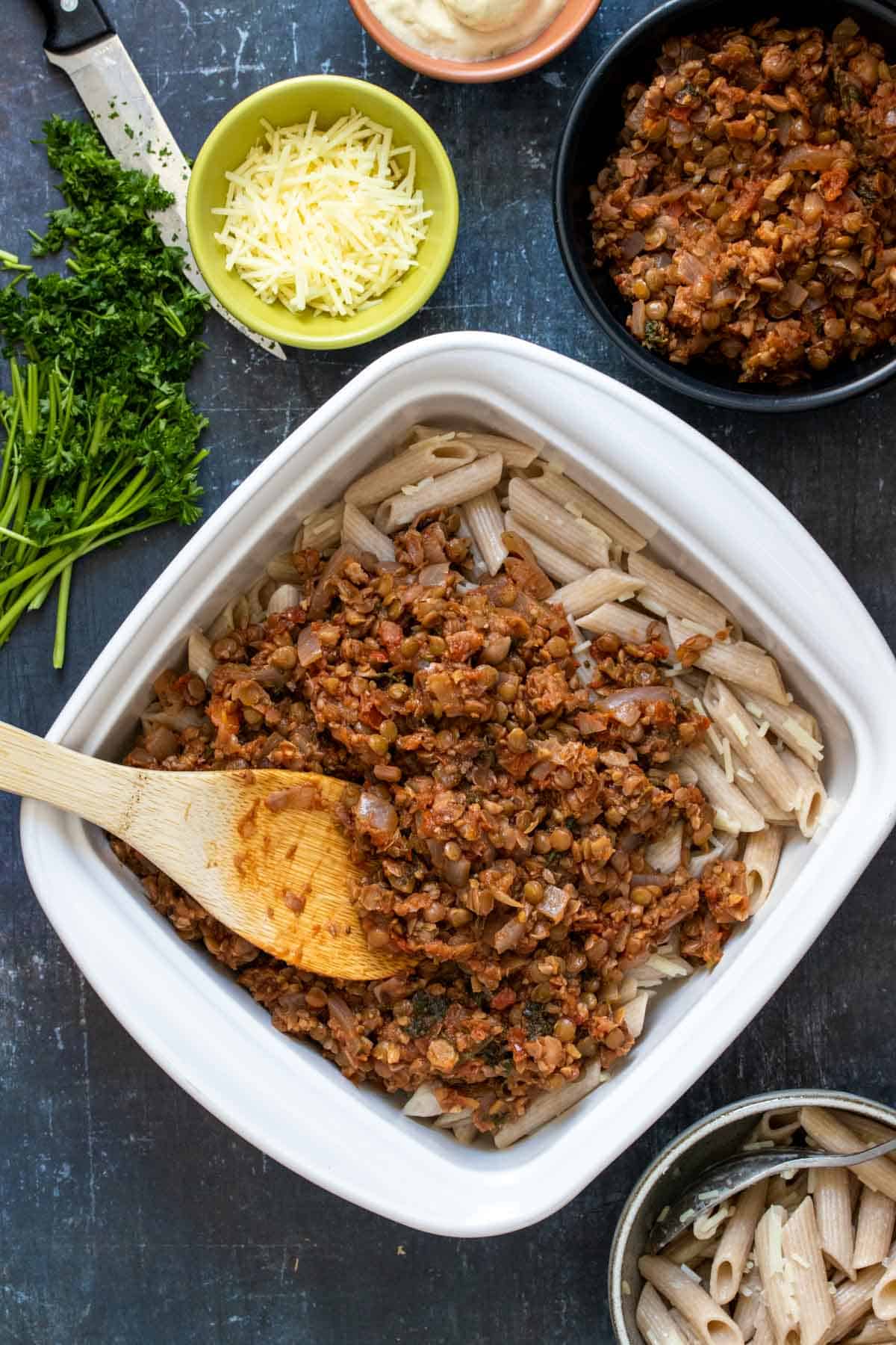 A wooden spoon spreading a meaty looking tomato based mixture on top of pasta in a white baking dish