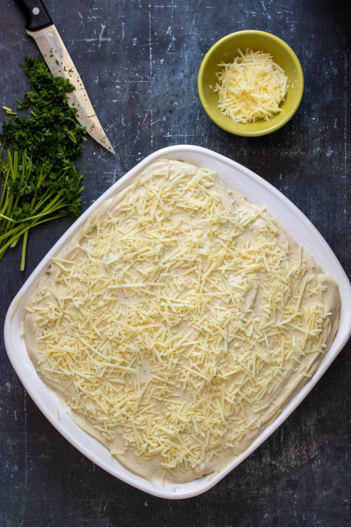 Top view of a white baking dish with Bechamel topped with Parmesan next to a bowl of Parmesan and chopped parsley