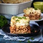 A black plate on a light napkin with a piece of pastitsio on it and more pastitsio in the background