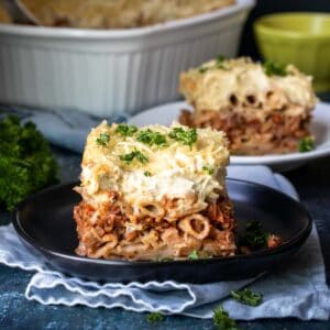 A black plate on a light napkin with a piece of pastitsio on it and more pastitsio in the background