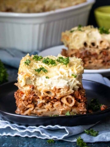 A black plate on a light napkin with a piece of pastitsio on it and more pastitsio in the background