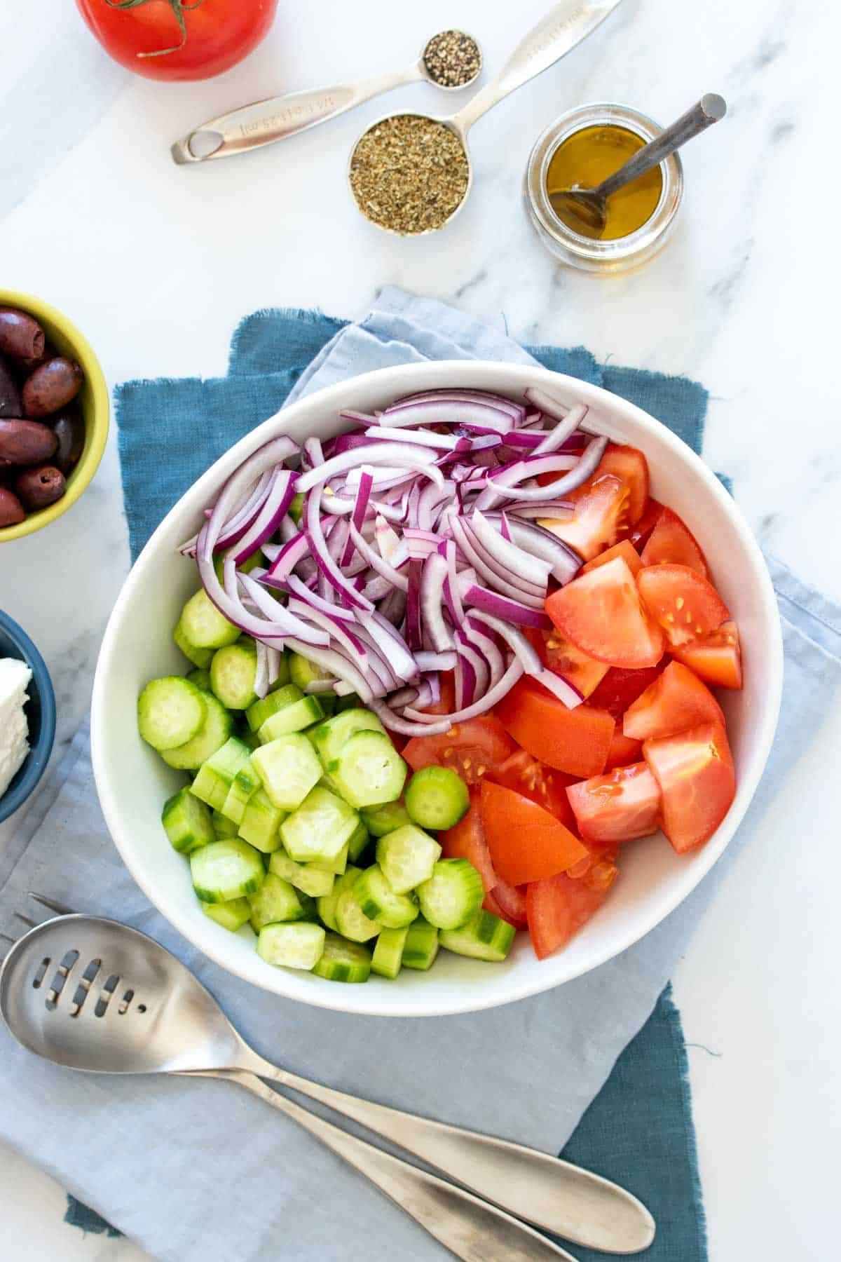 A large white bowl with cut tomatoes, cucumbers and onions inside on blue towels on a marble surface