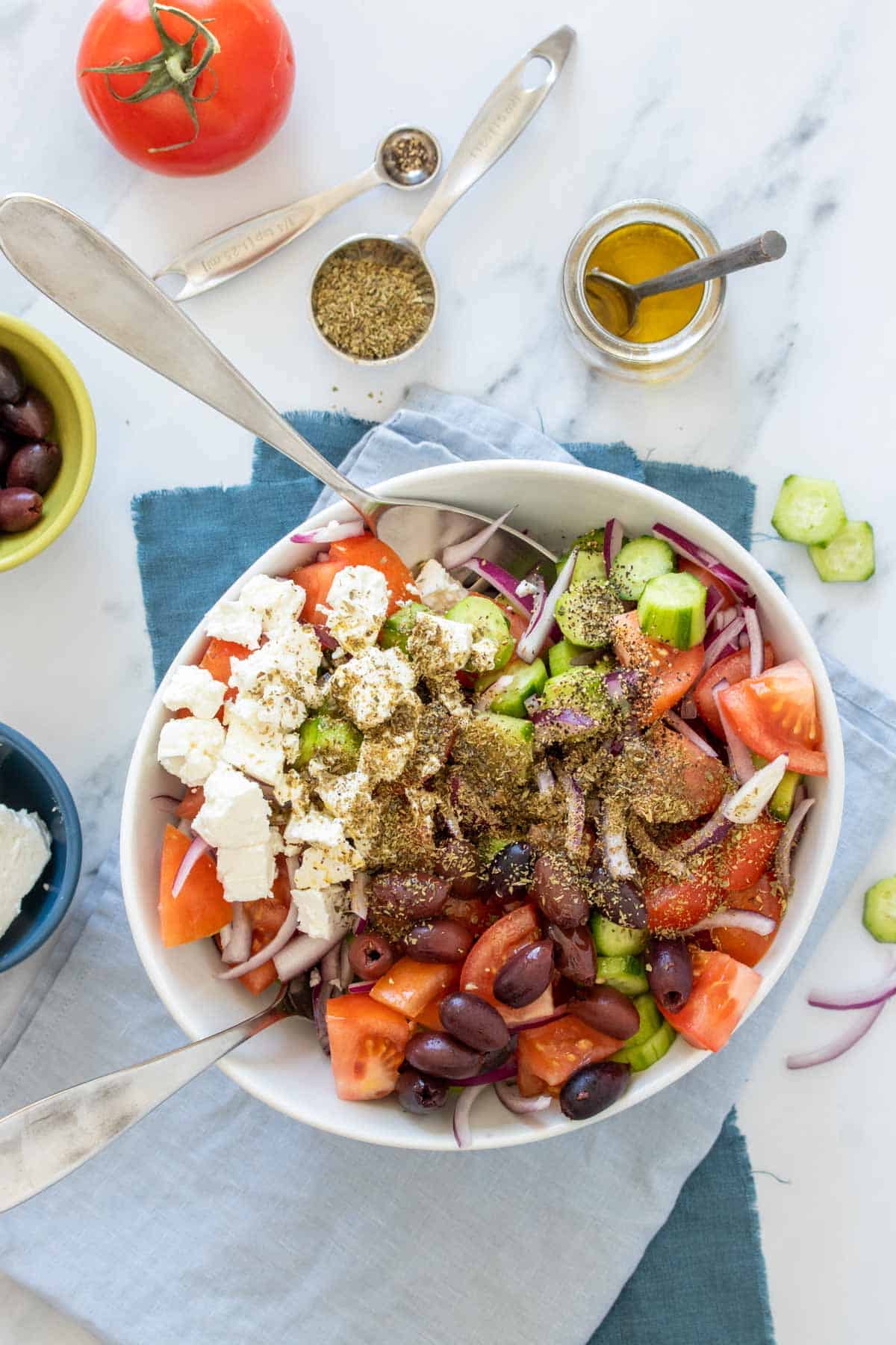 Two large spoons mixing a cucumber, tomato and red onion salad with feta, olives and oregano