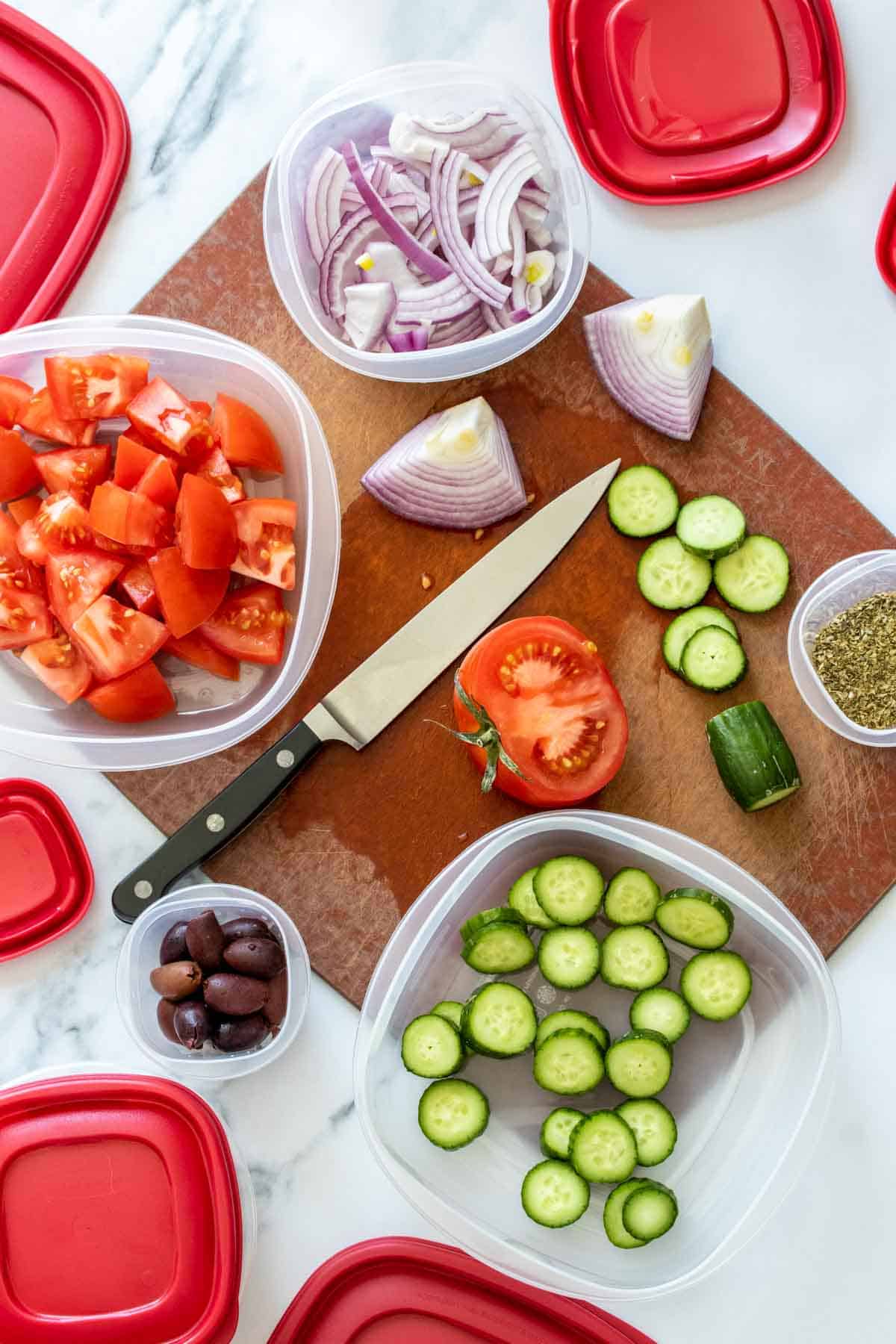 Top view of tomatoes, cucumbers and red onion being cut and put into plastic containers with red lids