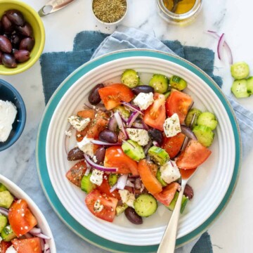 A white bowl on a turquoise plate with a Greek salad in it surrounded by the ingredients