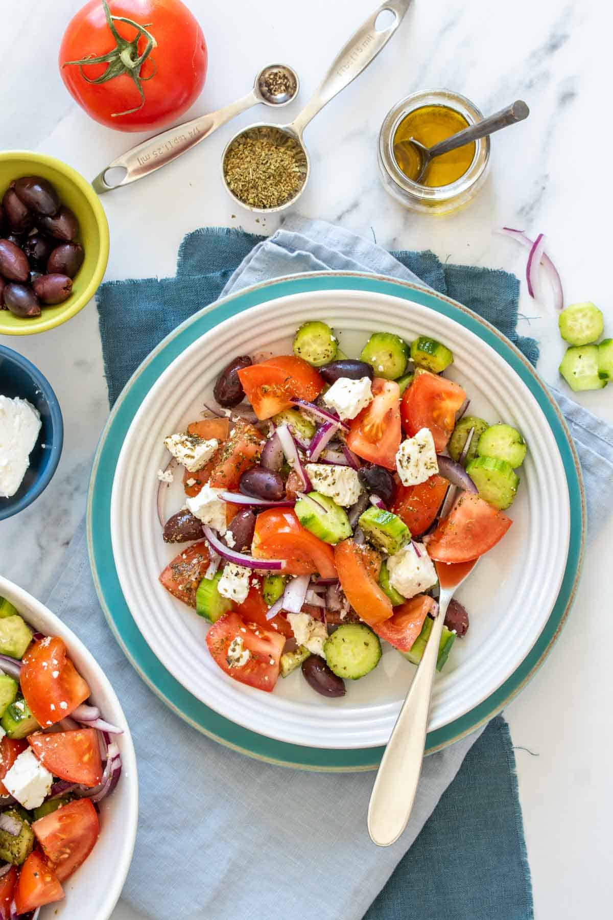A white bowl on a turquoise plate with a Greek salad in it surrounded by the ingredients