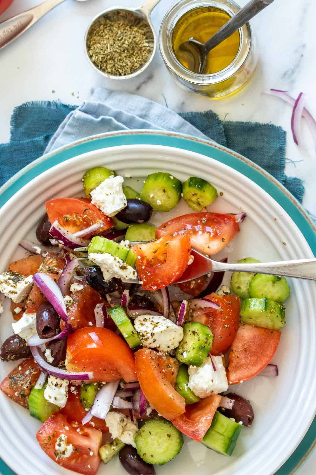 A fork getting a bite of Greek salad from a white bowl thats on a blue towel