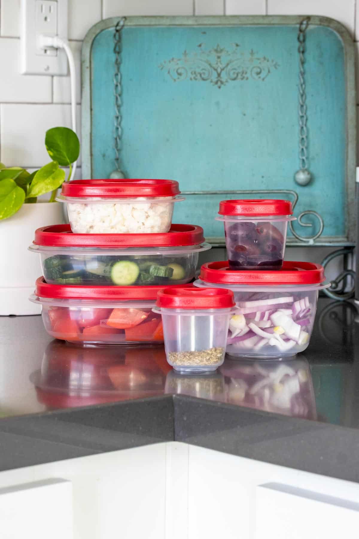 A stack of plastic Tupperware with red lids filled with ingredients for Greek salad stacked up on a counter