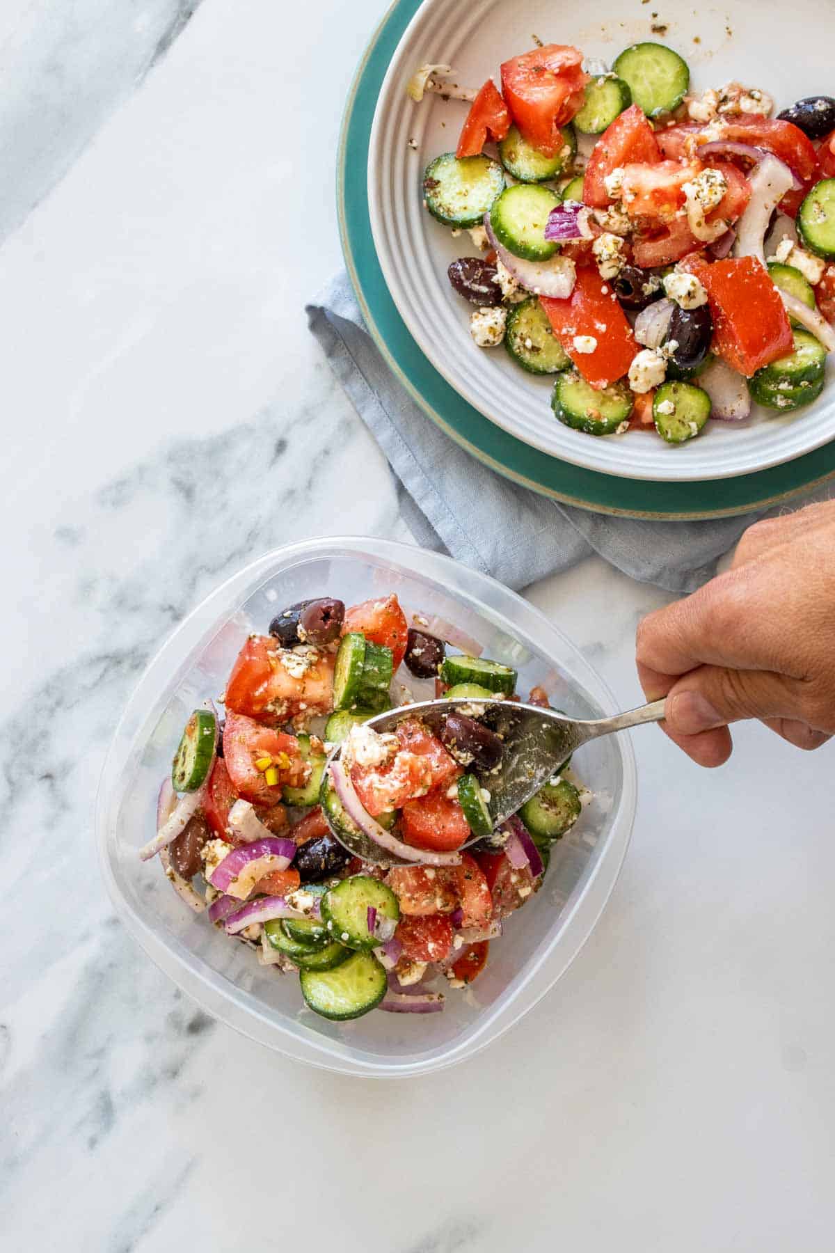 A hand scooping leftover Greek salad into a plastic container from a white bowl