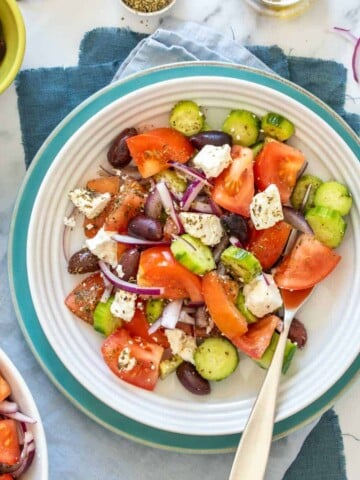 A Greek salad in a white shallow bowl that is sitting on a turquoise plate on towels on a marble surface