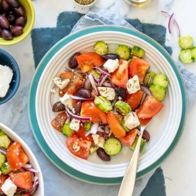 A Greek salad in a white shallow bowl that is sitting on a turquoise plate on towels on a marble surface