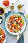 A white bowl of Greek salad with ingredients for is surrounding the bowl