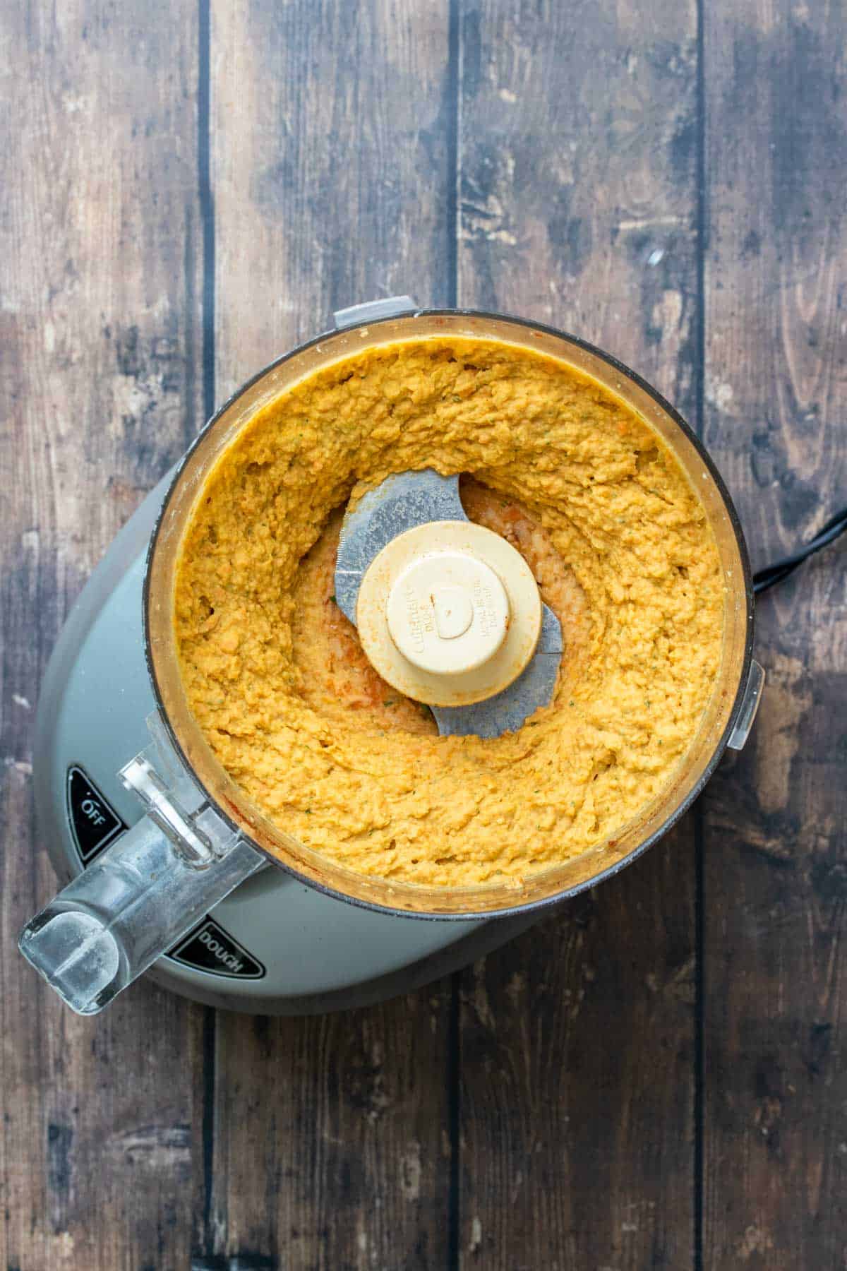 Top view of a food processor on a wooden surface with an orange colored hummus inside