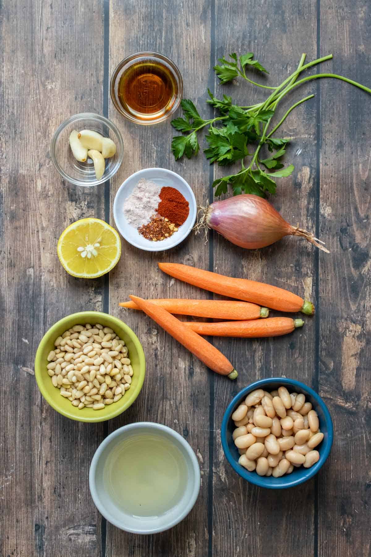 Ingredients on a wooden surface and in bowls that you would use to make a roasted carrot hummus with pine nuts and shallots