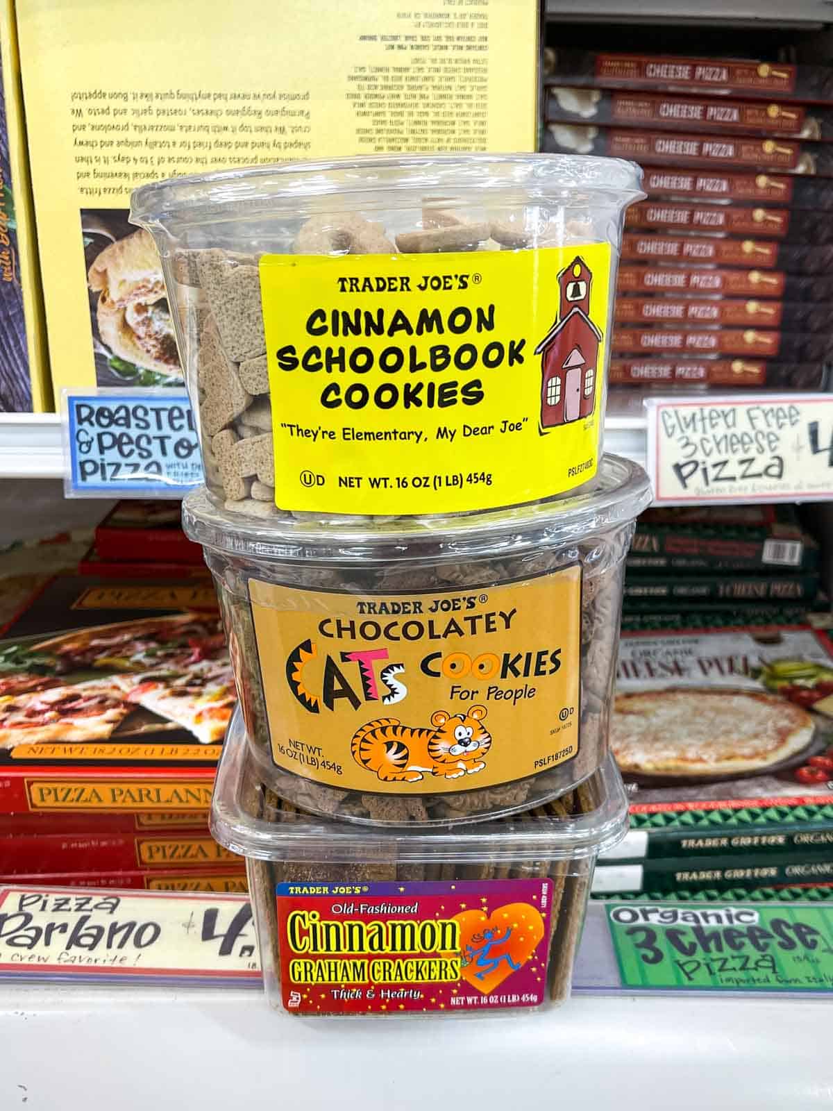 A pile of three plastic containers with different types of cookies sitting on the edge of a freezer grocery store shelf
