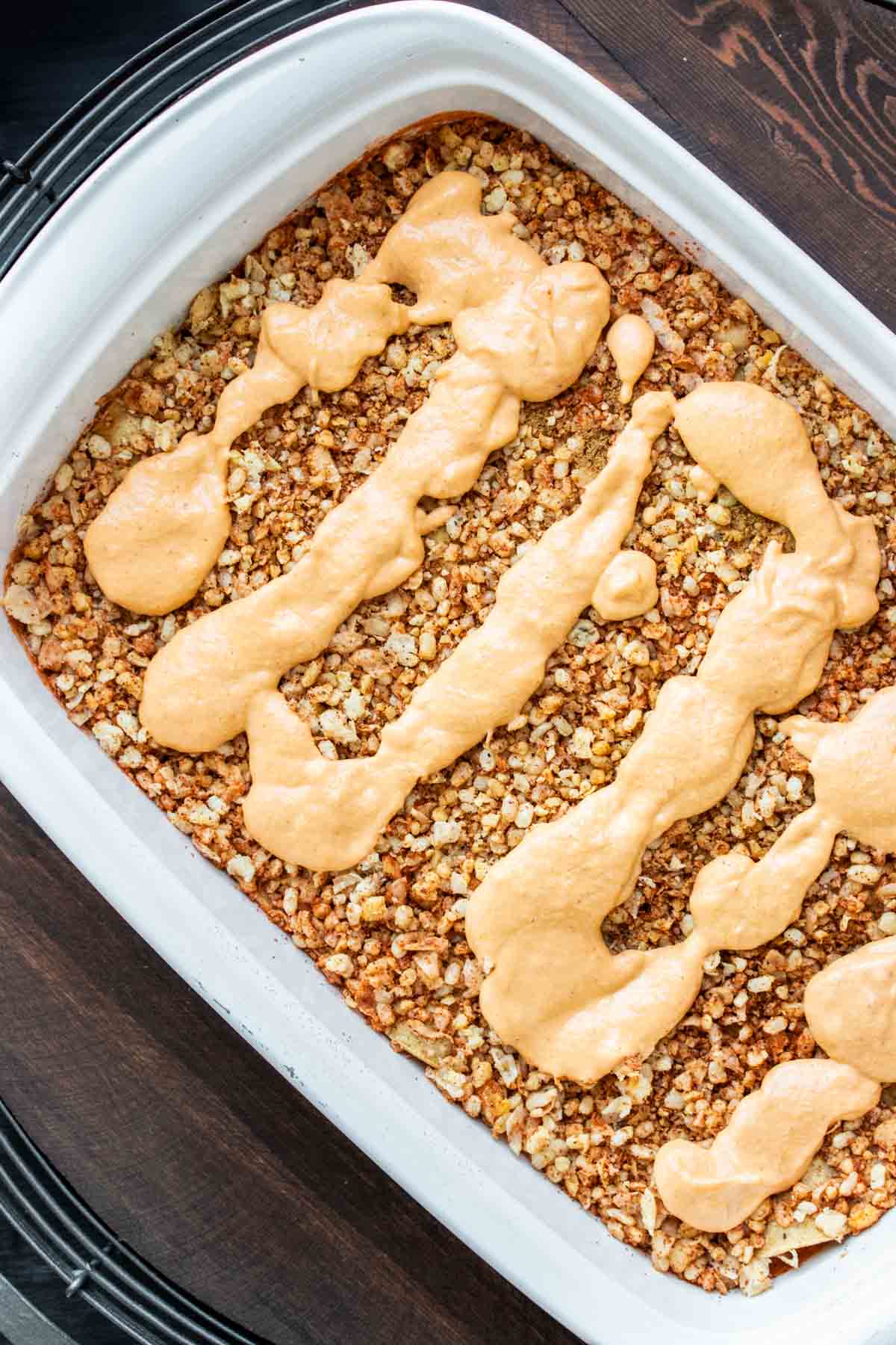 A baking dish with a meaty crumble topped with lines of a cheesy sauce in a white baking dish.