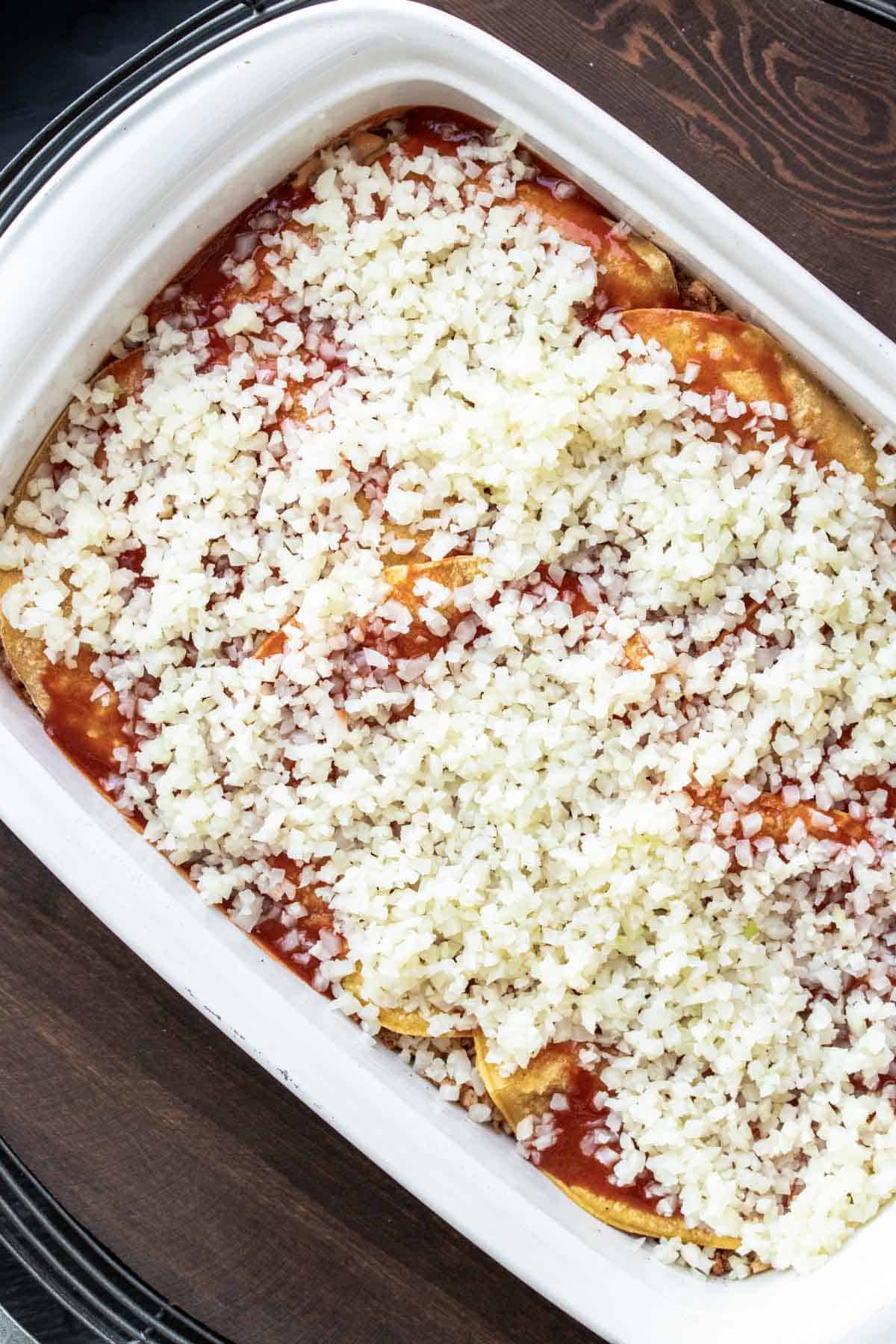 Top view of a white baking dish with layers of tortillas, enchilada sauce and riced cauliflower on top.