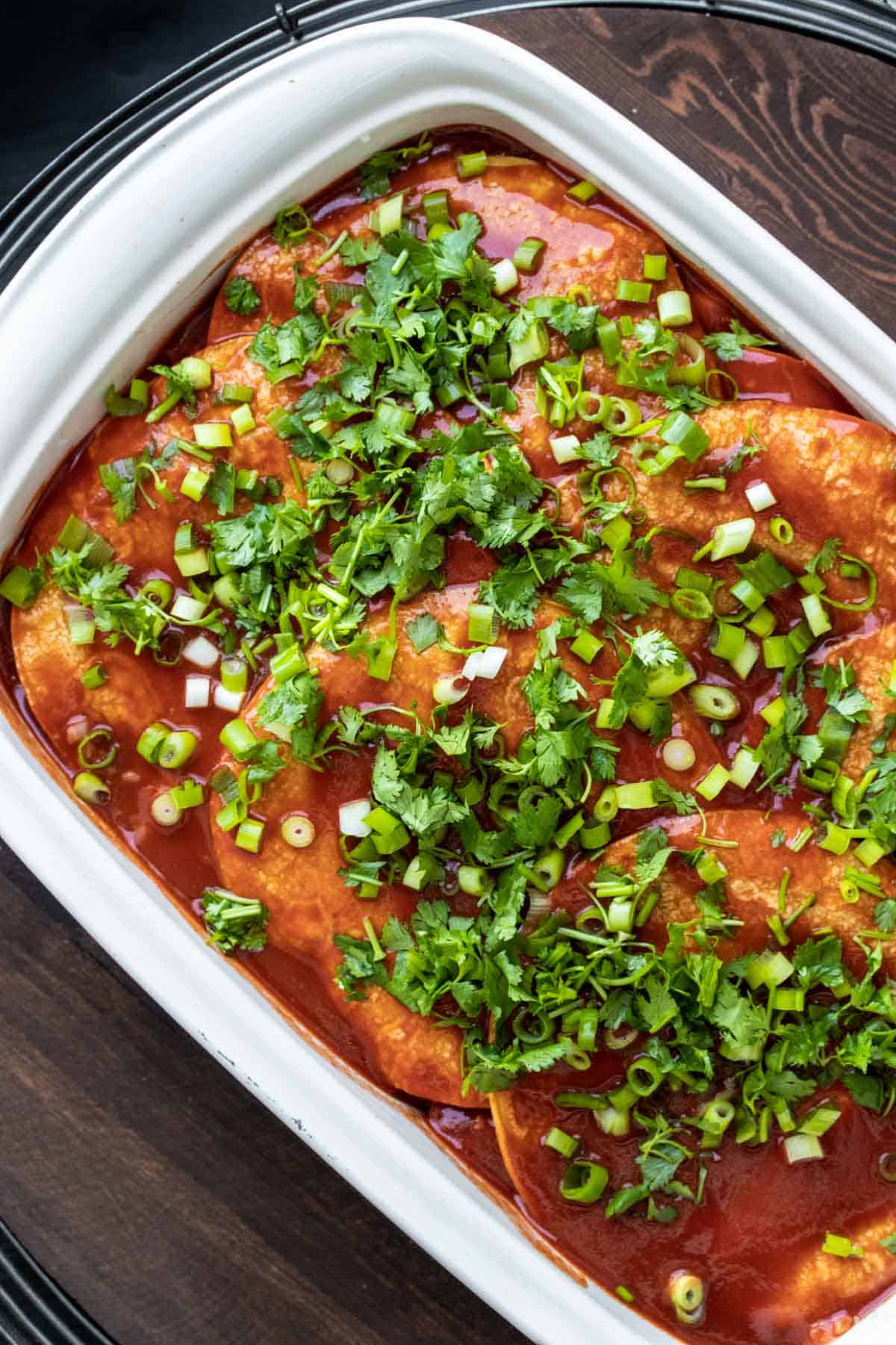 Top view of layered enchiladas in a white baking dish with red sauce and green onions.