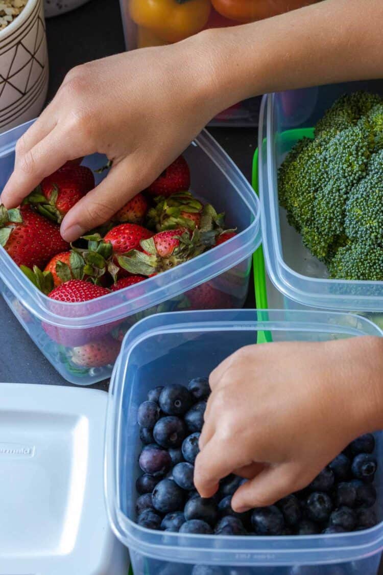 Hands grabbing strawberries and blueberries from plastic containers on a kitchen counter.