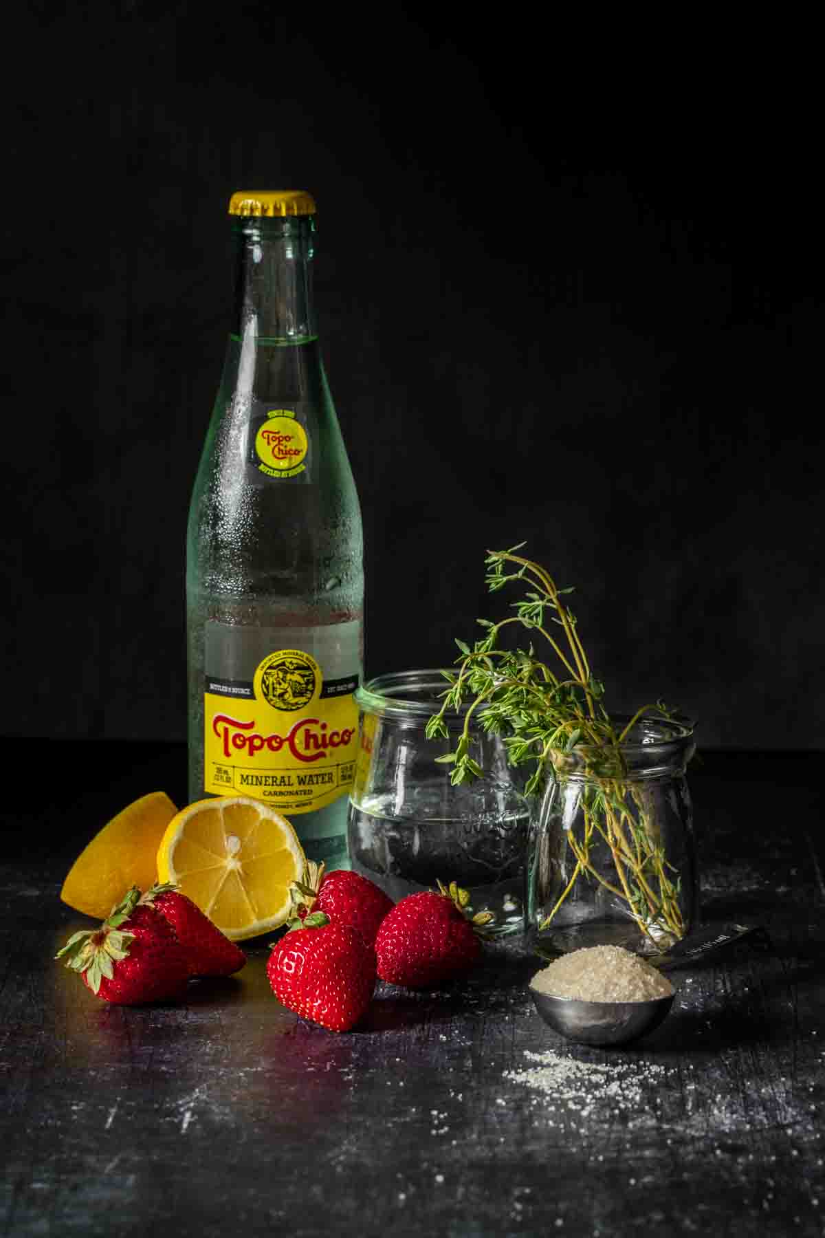 A bottle of Topo Chico next to strawberries, lemon wedges, a clear liquid, thyme and sugar in a spoon.