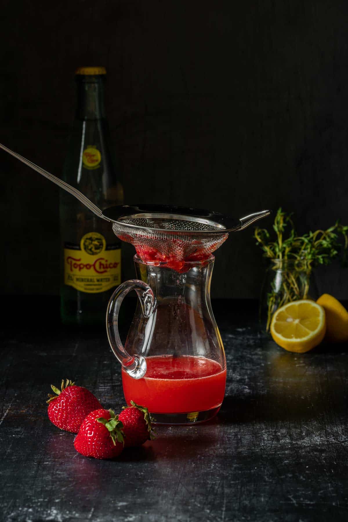 A strainer with mashed strawberries over a small glass pitcher with red juice.