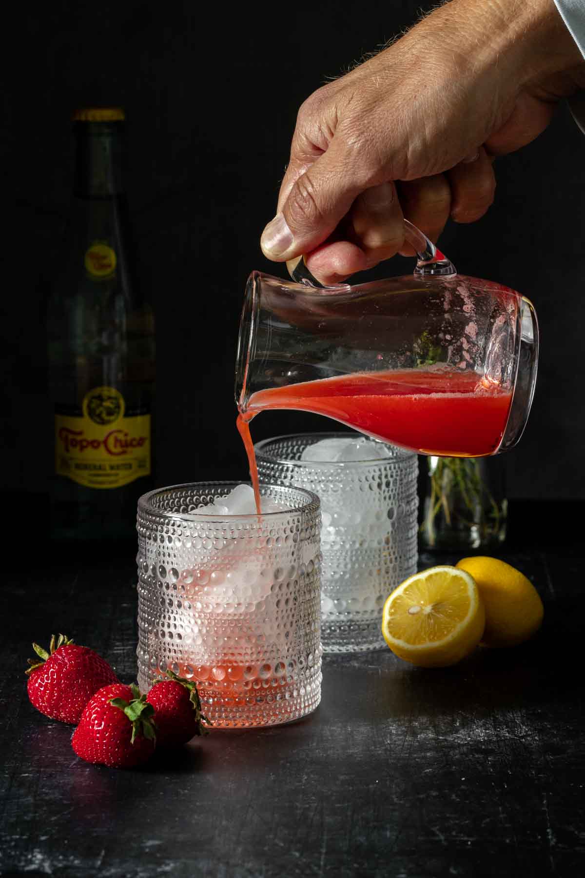 A hand pouring a red liquid into a textured cocktail glass with ice in it.