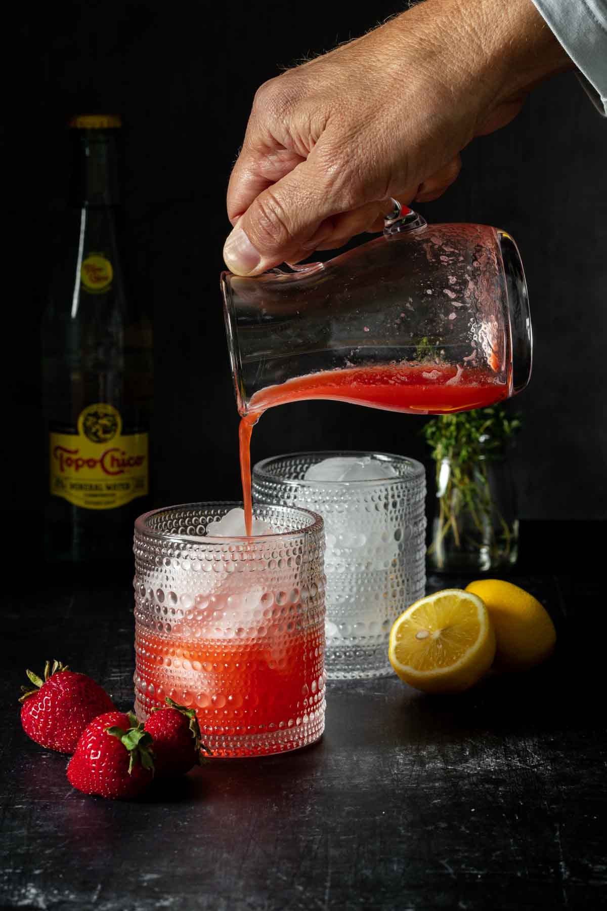 A hand filling up a cocktail glass with a red liquid over ice.
