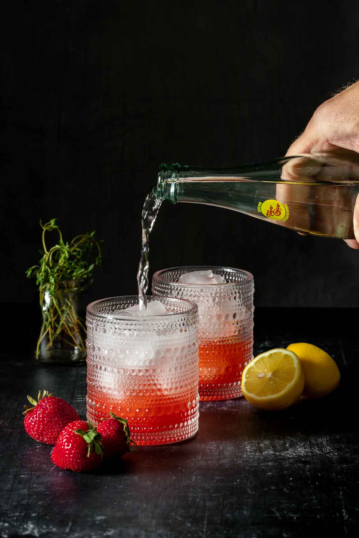 A hand pouring sparkling water from a glass bottle into a cocktail glass with ice and a red liquid.
