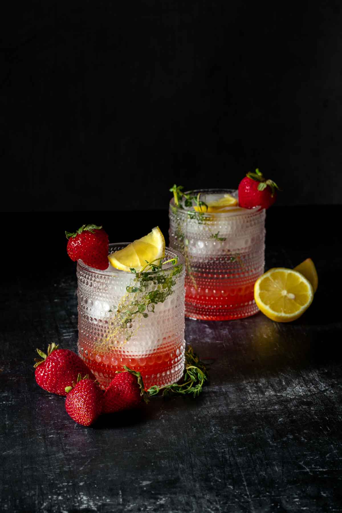 Two cocktail glasses with dots on them filled with a red liquid and ice and topped with lemon and thyme with a strawberry on the rim.