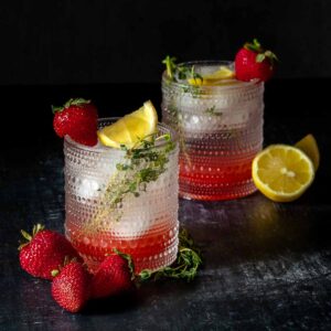 Two short glasses with a dots texture filled with a strawberry sparkling water cocktail and a lemon thyme garnish with a strawberry on the rim.