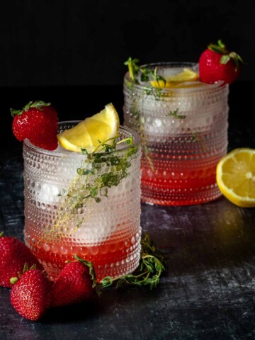 Two short glasses with a dots texture filled with a strawberry sparkling water cocktail and a lemon thyme garnish with a strawberry on the rim.