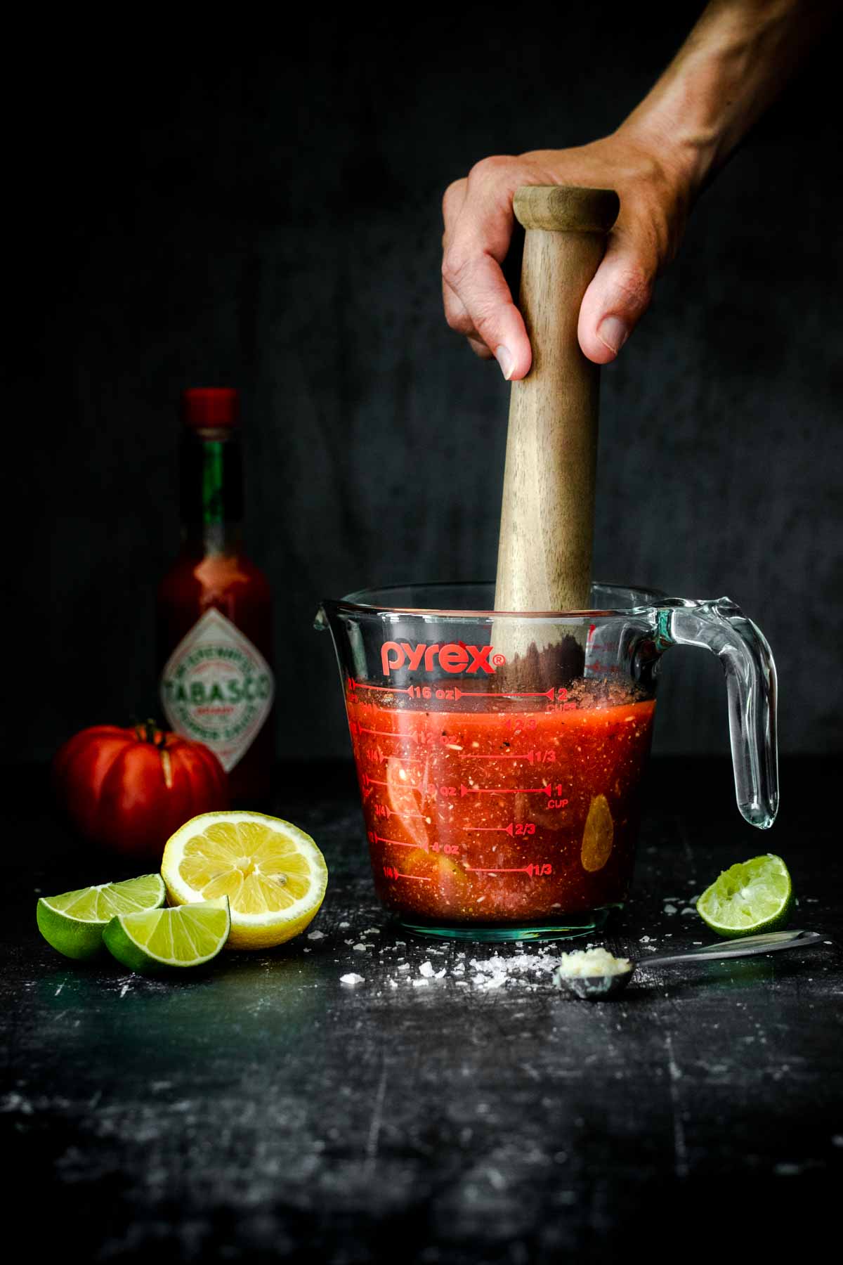 Hand using a wooden muddler to muddle a bloody Mary in a glass measuring cup.