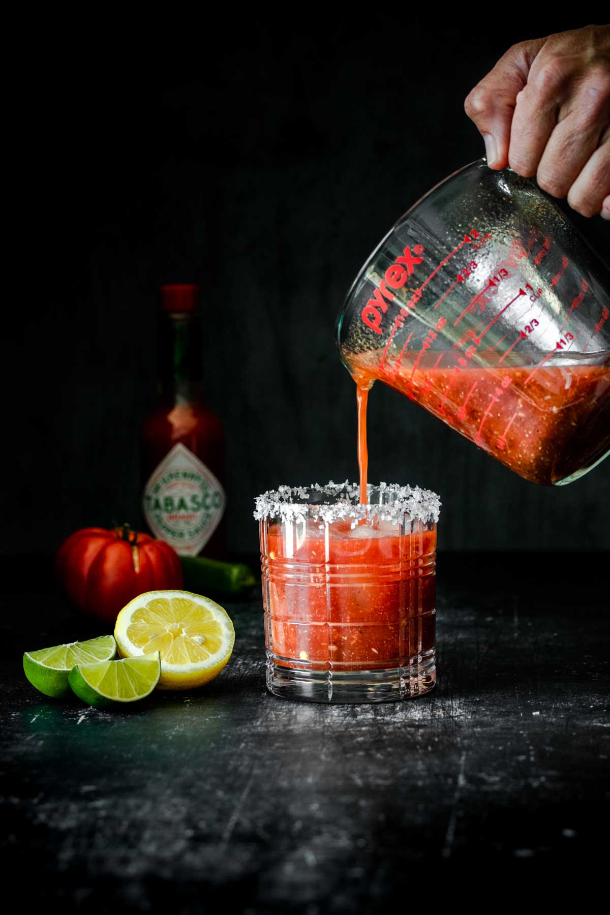 A hand filling a glass with ice with a bloody Mary mix from a measuring cup.
