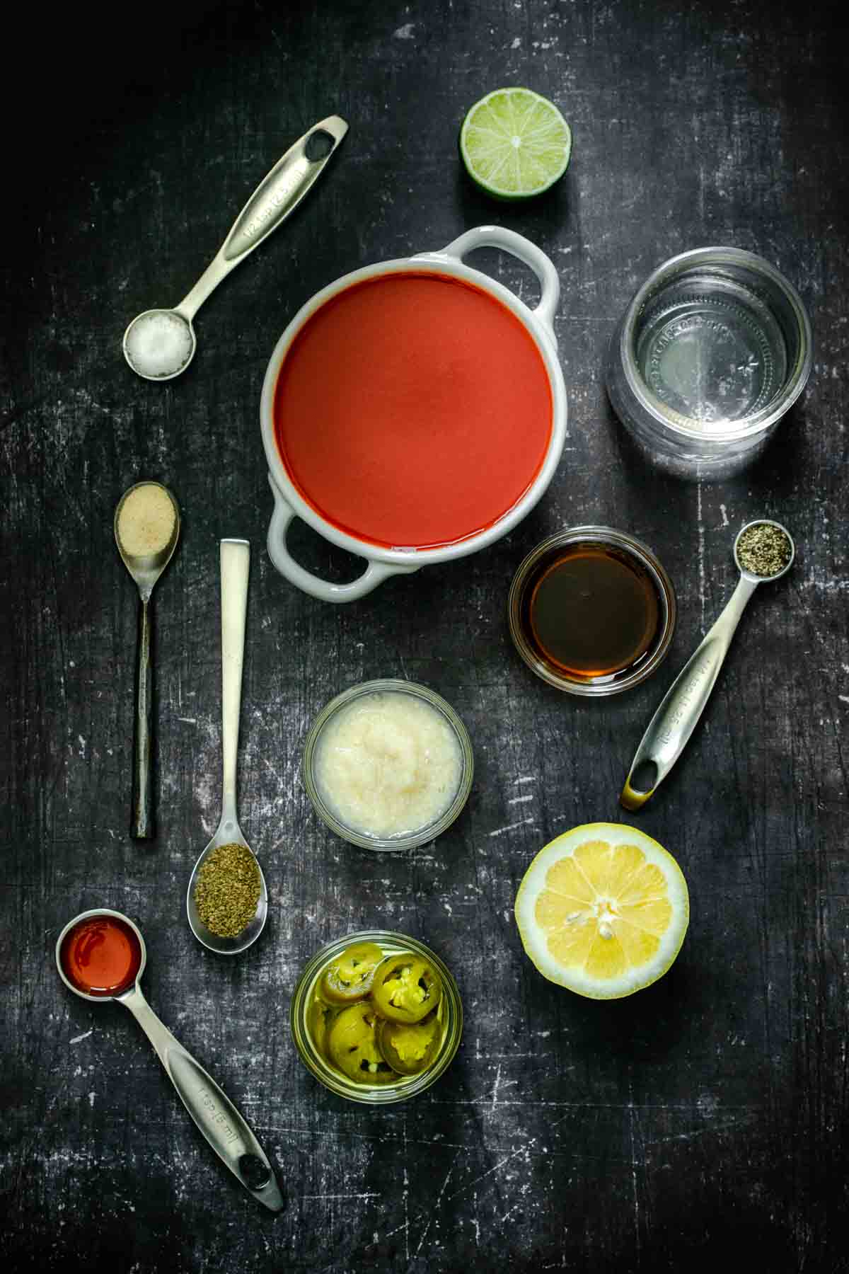 Top view of a dark surface with ingredients for a bloody Mary in bowls and spoons.