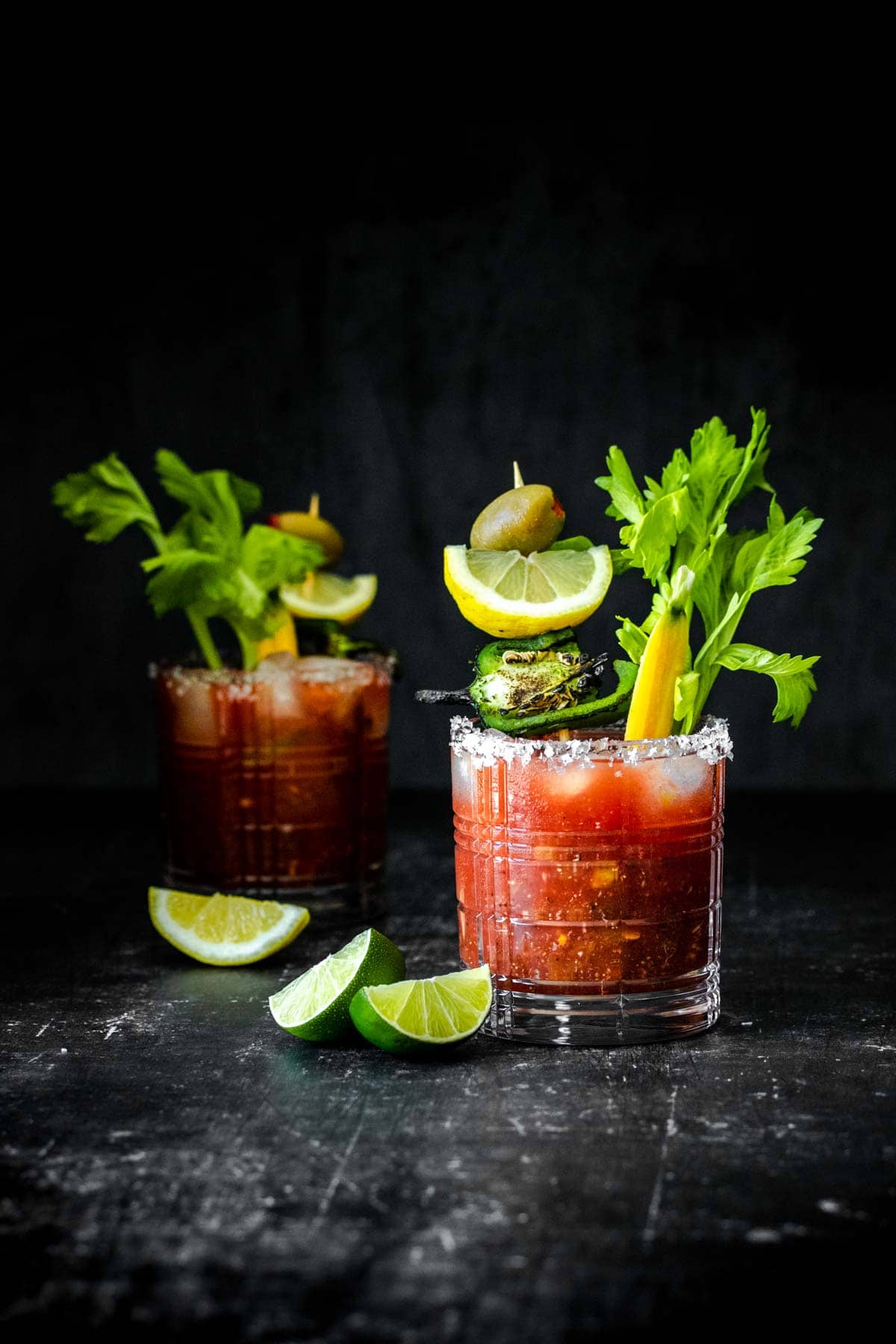 A lowball cocktail glass with ice and a bloody Mary in it filled with veggies in front of another one.