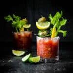 Two glasses filled with a bloody Mary cocktail and veggie garnishes with one in the background.