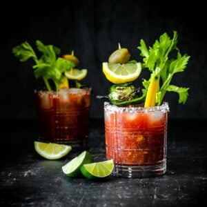 Two glasses filled with a bloody Mary cocktail and veggie garnishes with one in the background.