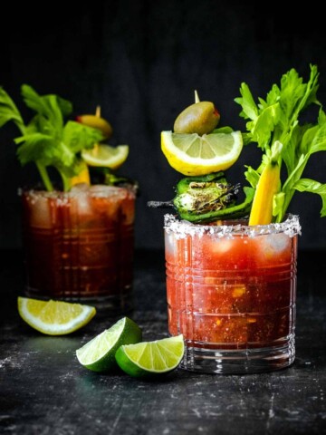 Two glasses filled with a bloody Mary cocktail and veggie garnishes with one in the background.