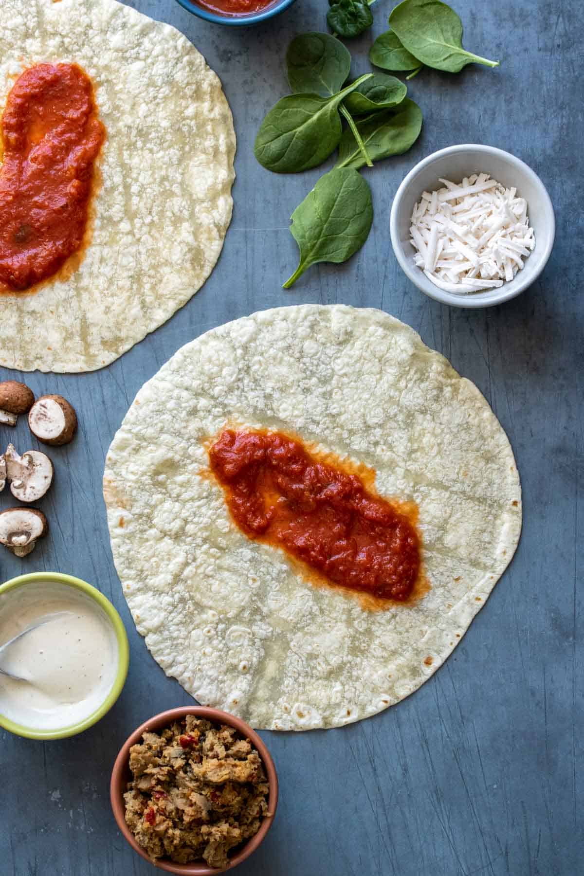 A tortilla with pizza sauce in the center on a slate surface surrounded by pizza ingredients