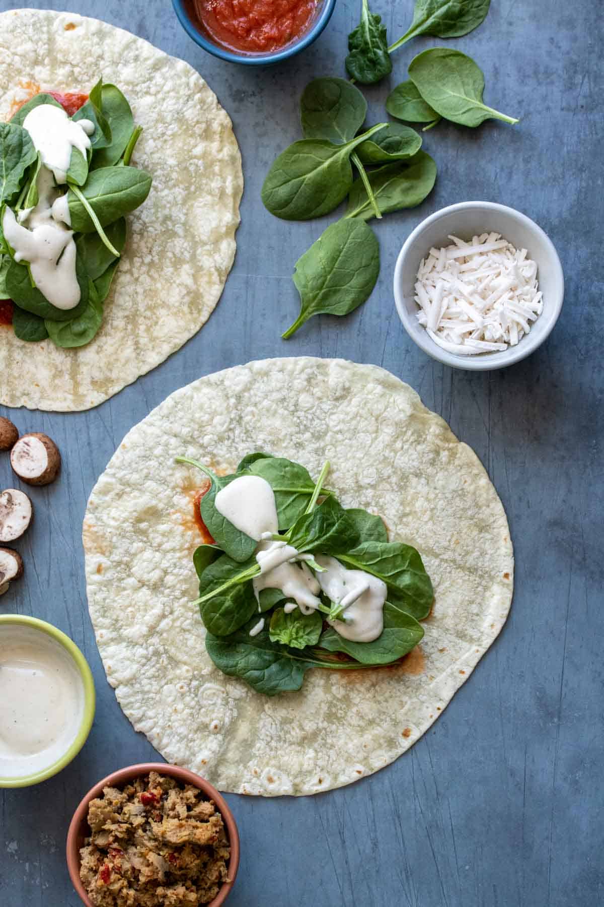 A tortilla with spinach and white sauce in the center on a slate surface and other pizza ingredients around it