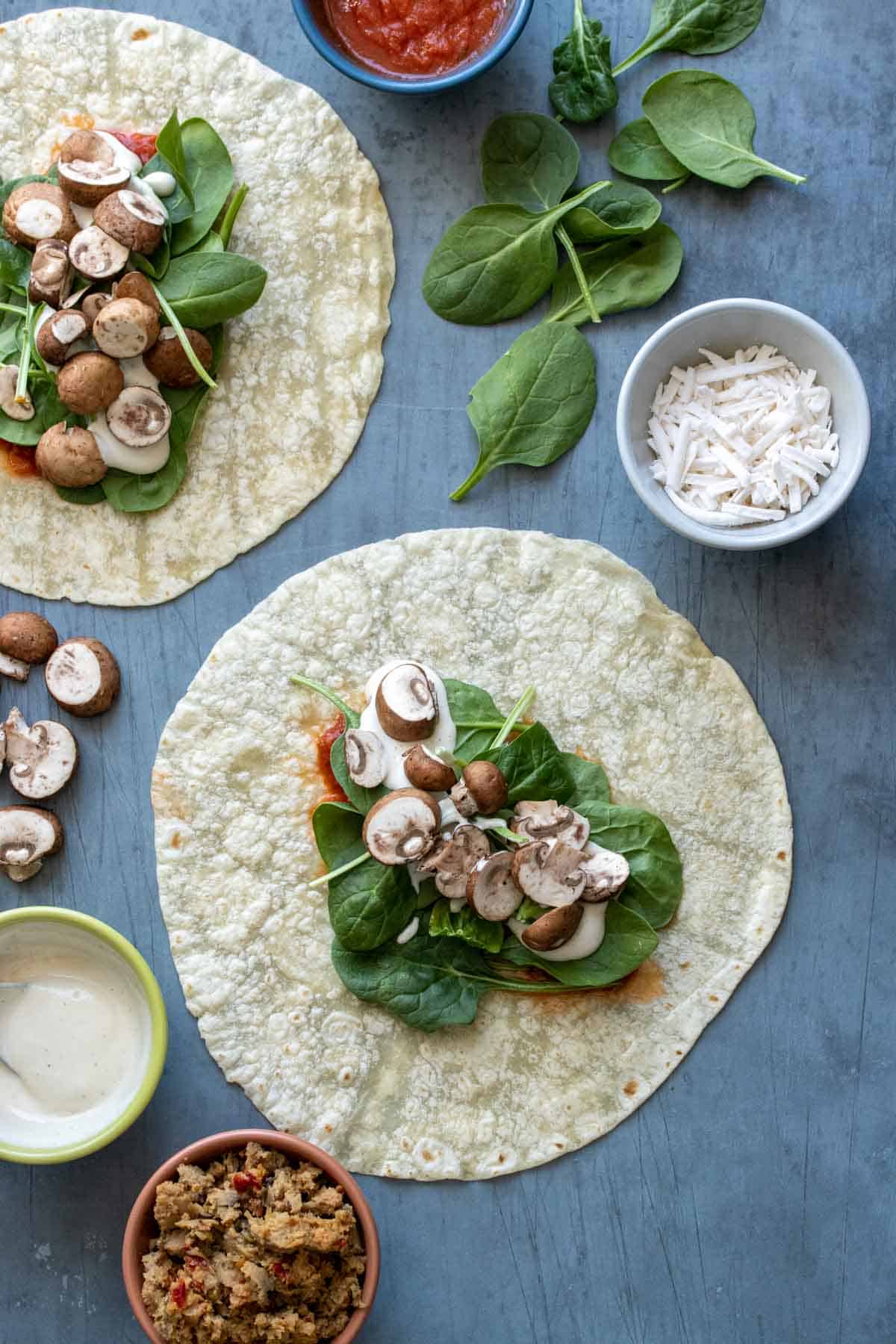 A tortilla on a grey surface with spinach, sauce and mushrooms on it and other pizza ingredients around