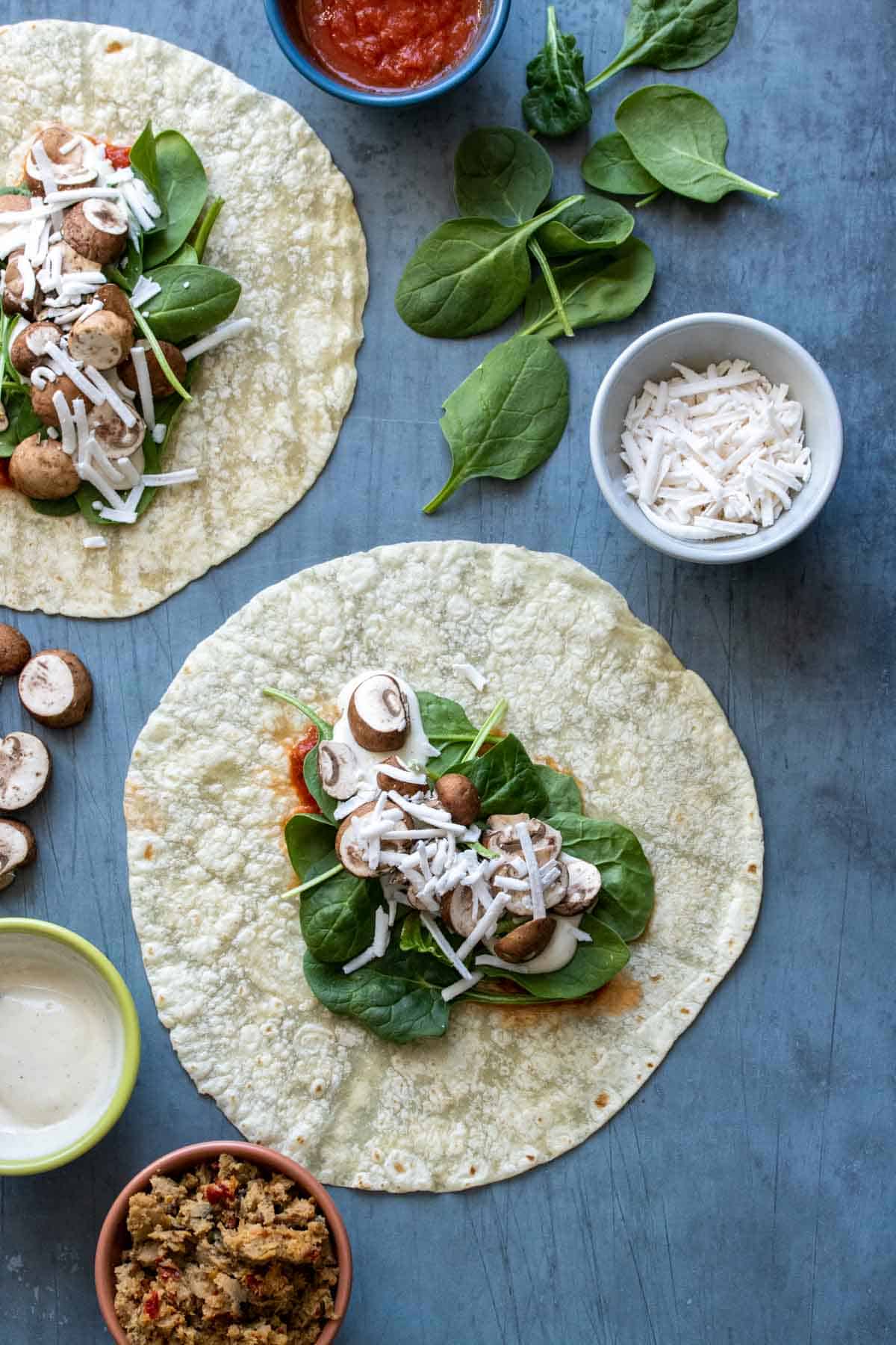 A tortilla covered in spinach, mushrooms, sauce and cheese on a slate surface and pizza ingredients around it