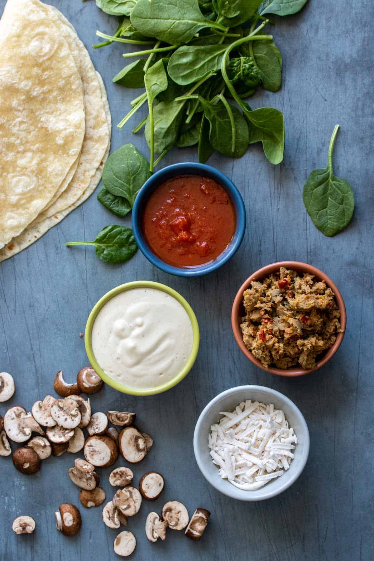 A grey surface with mushrooms, spinach, tortillas on it and sausage, alfredo, pizza sauce and cheese in bowls