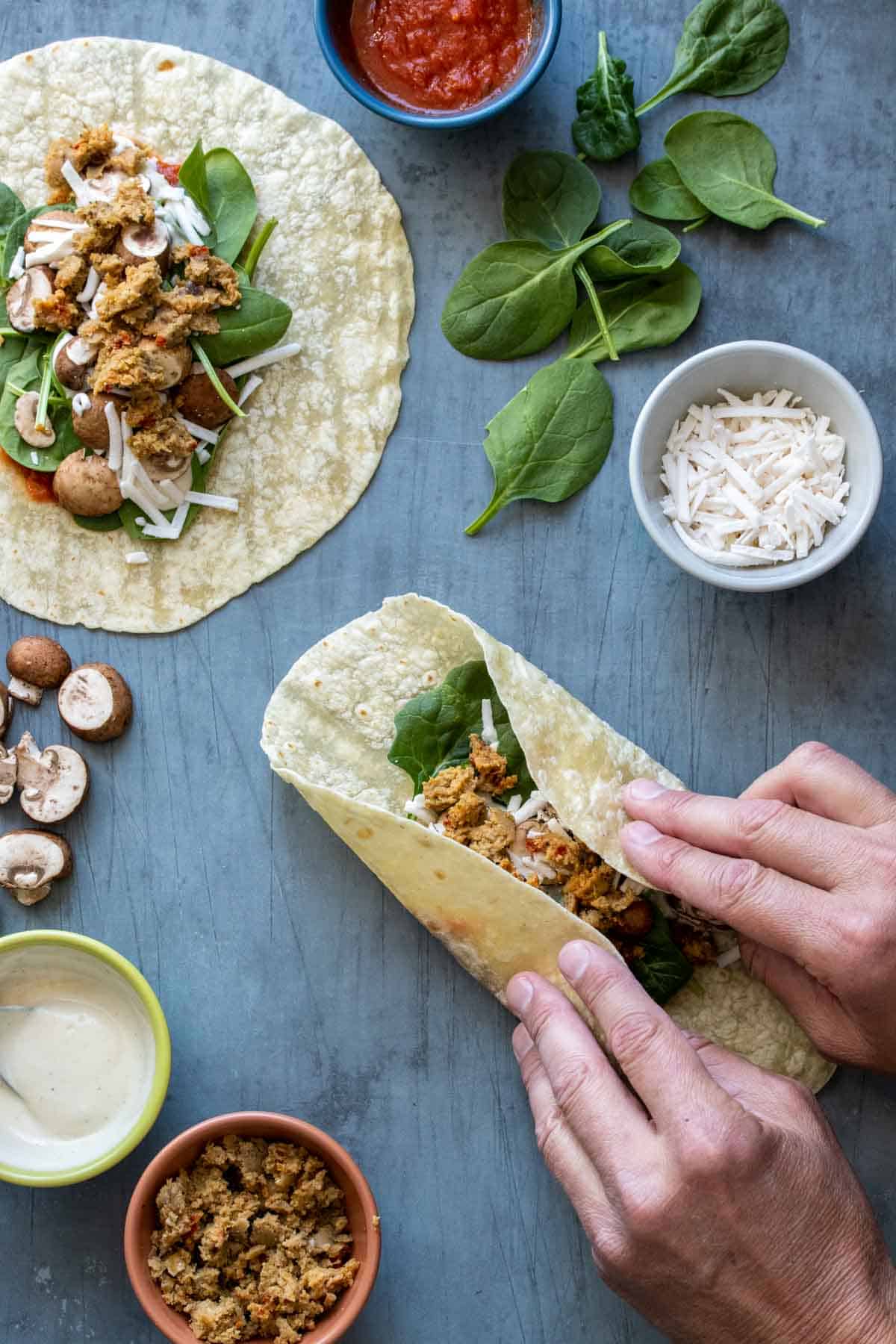 Two hands folding the sides of a tortilla with ingredients inside towards the center with another open faced tortilla next to it